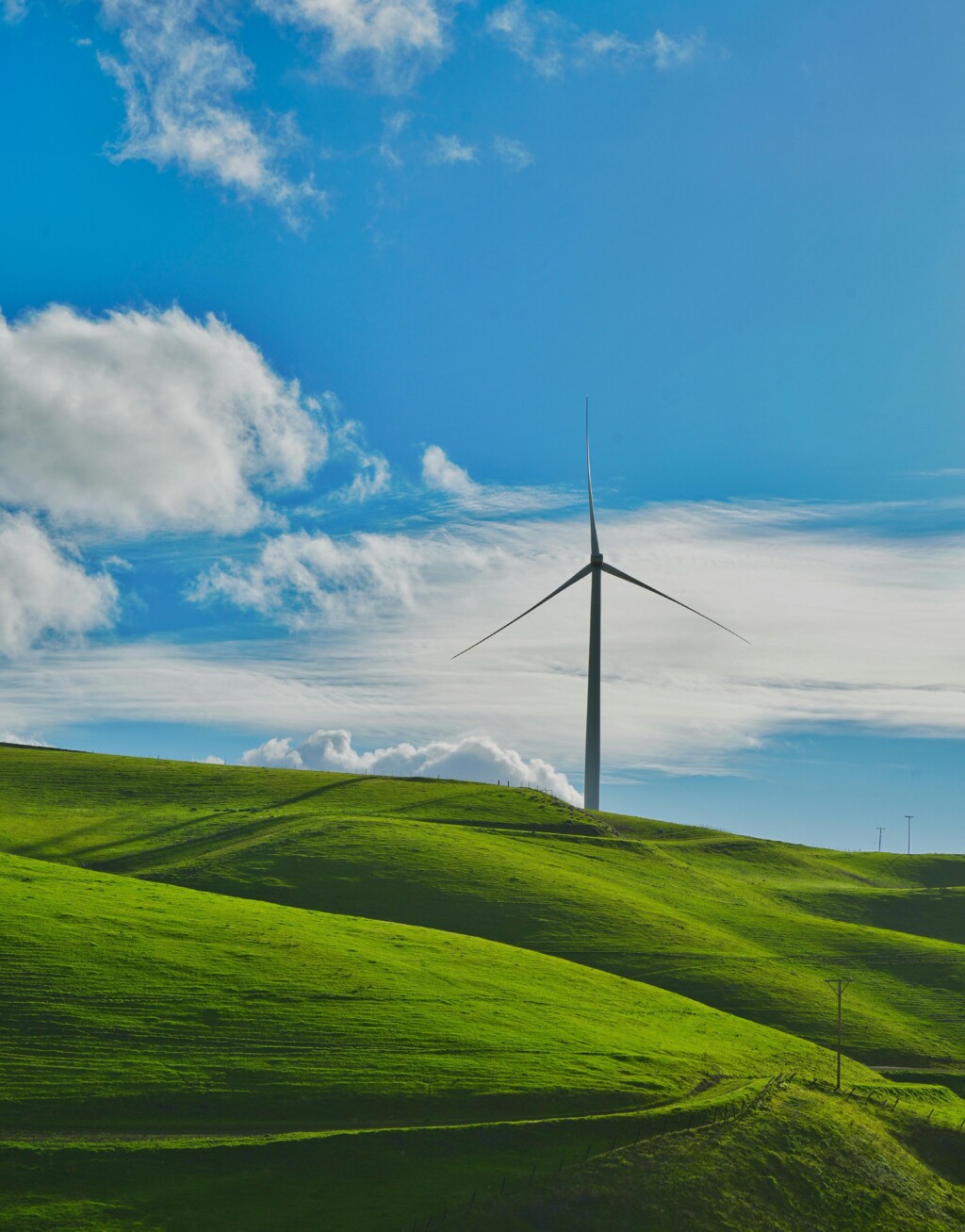 A wind turbine stands prominently against rolling green hills and a bright blue sky with clouds.