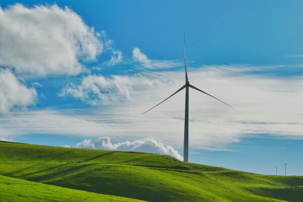 A wind turbine stands prominently against rolling green hills and a bright blue sky with clouds.