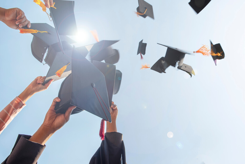 Graduates arms throwing graduation caps into the blue sky.