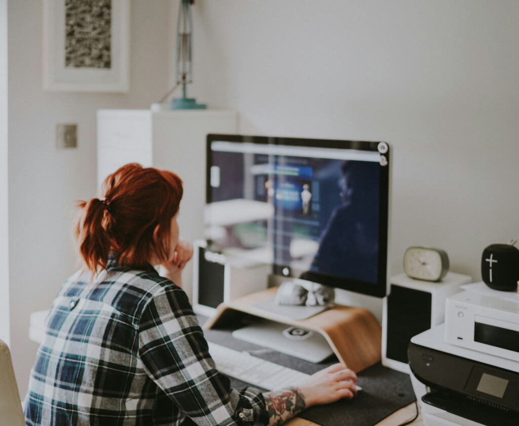 A woman with red hair and a plaid shirt works at a desk in her home office