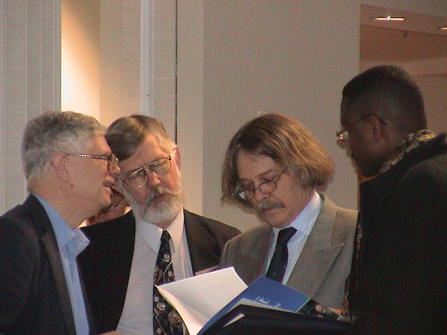 Nicolas is standing between three other professors and they are looking at the program of a symposium which he is holding.