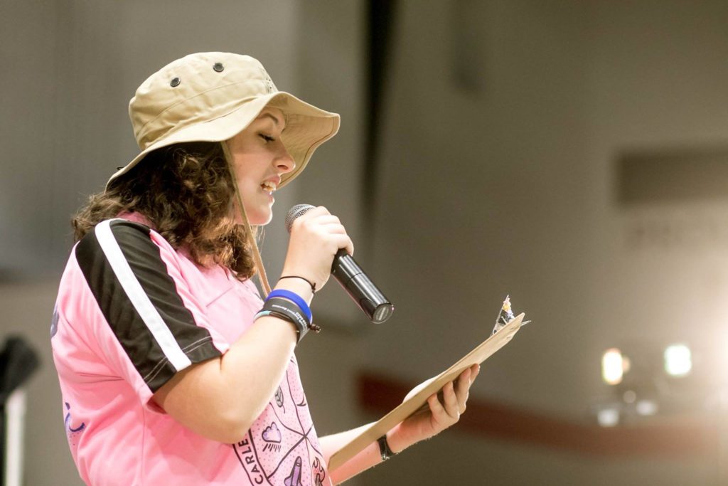 Emma wearing her Sprosh shirt and hat reading off a clipboard into a microphone