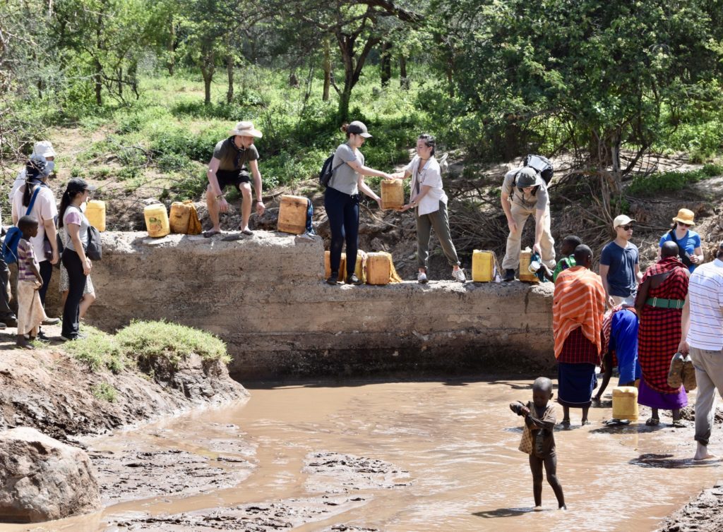 Carleton and Sprott students collecting water with local women and children at muddy water source in Longido, Tanzania.