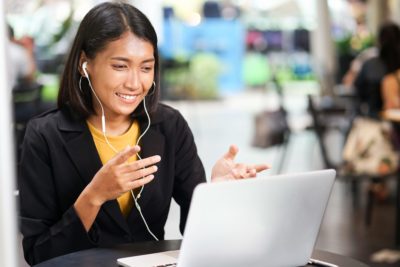 Picture of a woman talking into her laptop with earphones on. She is gesturing with both hands.