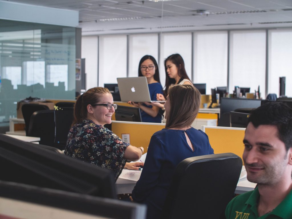 a female student discusses with her mentor in a library. 2 female students at a computer are in the background and a male student on a computer in the foreground.