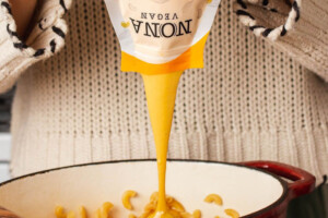 A person is pouring NONA Vegan sauce into a bowl of macaroni.