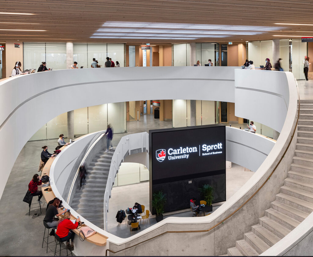 View from he fourth level looking down into open atrium on the ground level. Large media screen in atrium visible. Students working along the counters that wind around the floors and staircases.