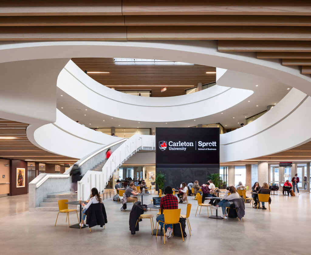 Nicol Building atrium on ground floor. People sitting at tables and interacting. Large media screen next to a large winding staircarse leading up to third level. Sunlight from skylight shining down on open atrium below.