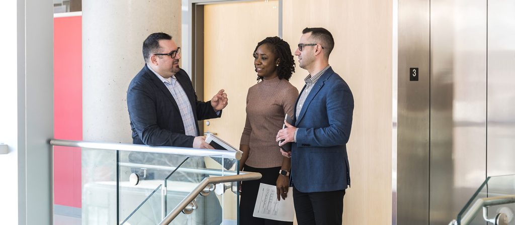 Group of three Professional MBA students engaged in a discussion near a staircase.