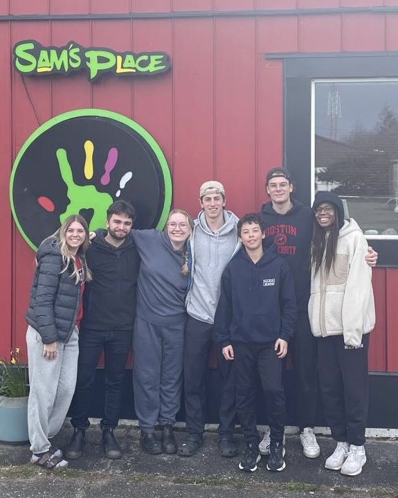 Six Carleton students and a younger child stand outside a red building that says Sam's Place