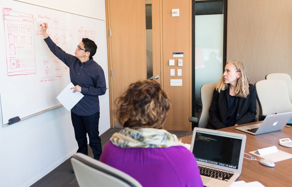 a man writing on a whiteboard with 2 women seated at a boardroom table