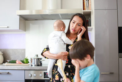 Mother talking on the phone with an upset expression, holding a baby and a small child nearby.